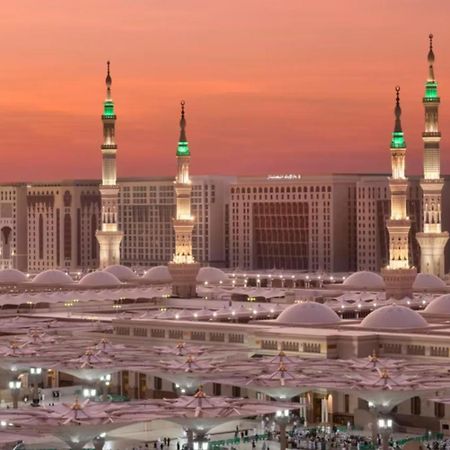 Intercontinental Dar Al Iman Madinah By Ihg Hotel Medina Buitenkant foto The photo depicts a scenic view of a mosque complex, likely during sunset, with several minarets illuminated and showcasing green lights. The architecture features domed roofs, and the setting includes a backdrop of buildings, all bathed in a warm, o