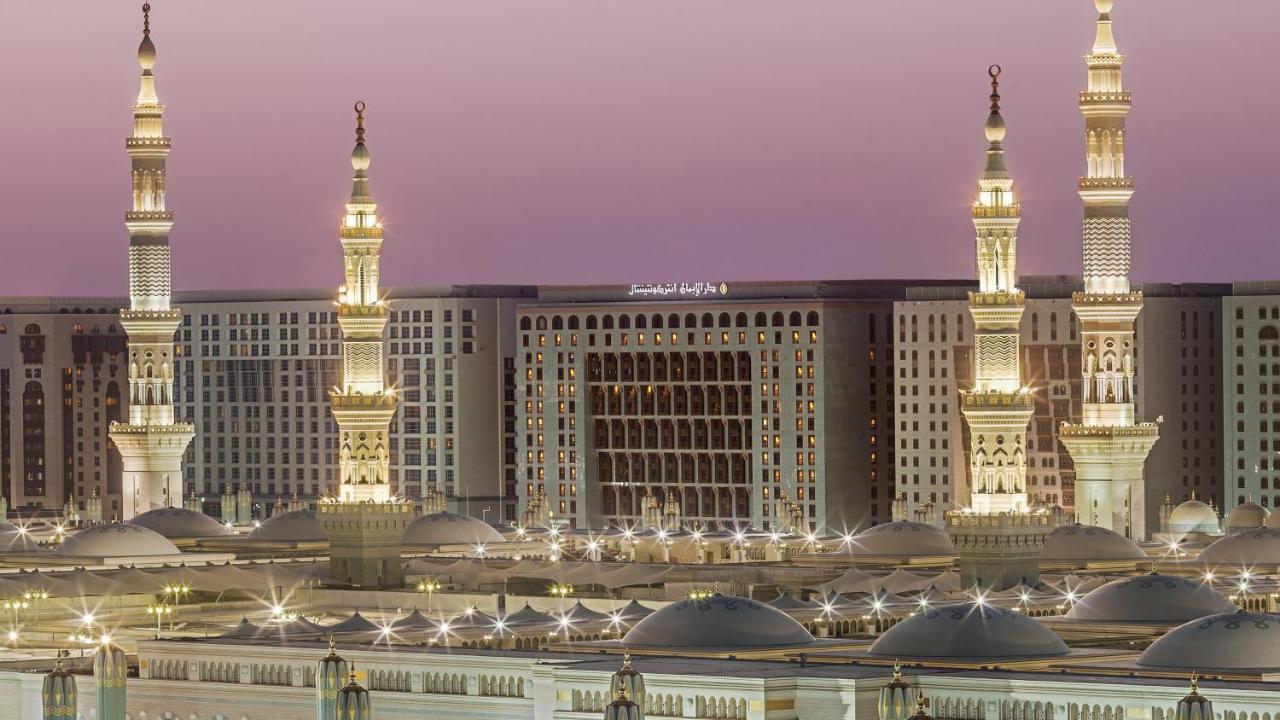 Intercontinental Dar Al Iman Madinah By Ihg Hotel Medina Buitenkant foto The photo depicts an architectural scene, likely from a mosque complex, featuring prominent minarets that are illuminated. The setting appears to be during twilight, with a soft gradient in the sky. In the background, a large building is visible, pos