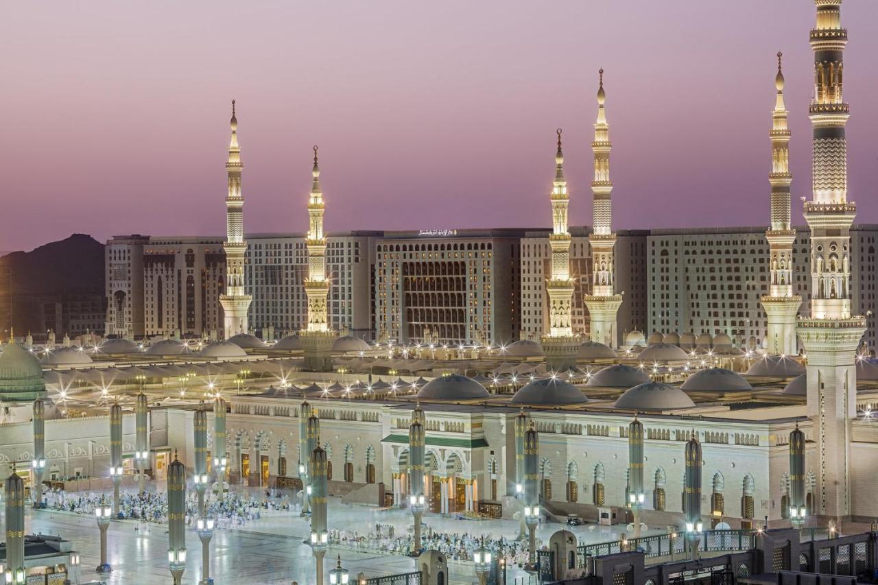 Intercontinental Dar Al Iman Madinah By Ihg Hotel Medina Buitenkant foto The photo shows a view of an expansive mosque complex, featuring several tall minarets that are illuminated against a twilight sky. The structure appears grand and is likely adorned with intricate architectural details. There are people gathered in t