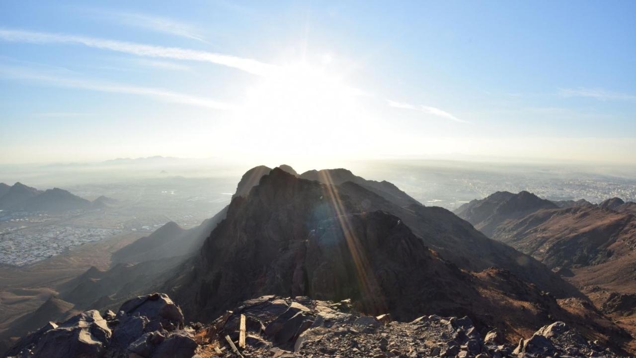 Intercontinental Dar Al Iman Madinah By Ihg Hotel Medina Buitenkant foto The photo shows a scenic view from a mountain top, with peaks silhouetted against the bright sun. The sun is positioned low in the sky, creating a warm glow and illuminating the landscape below. In the foreground, there are rocky terrains, while the 