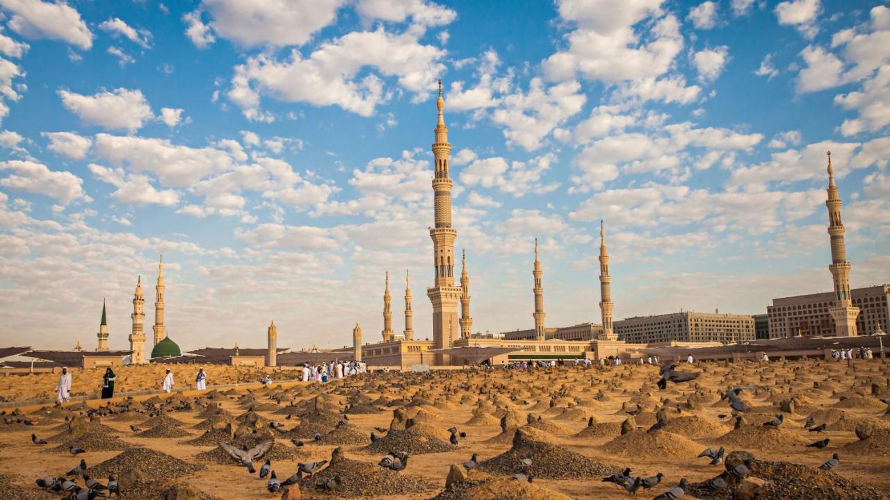 Intercontinental Dar Al Iman Madinah By Ihg Hotel Medina Buitenkant foto The photo shows a landscape that features a large mosque with a tall minaret surrounded by smaller minarets. In the foreground, there are numerous grave markers arranged in a desert-like setting. The sky is bright with scattered clouds, and there are