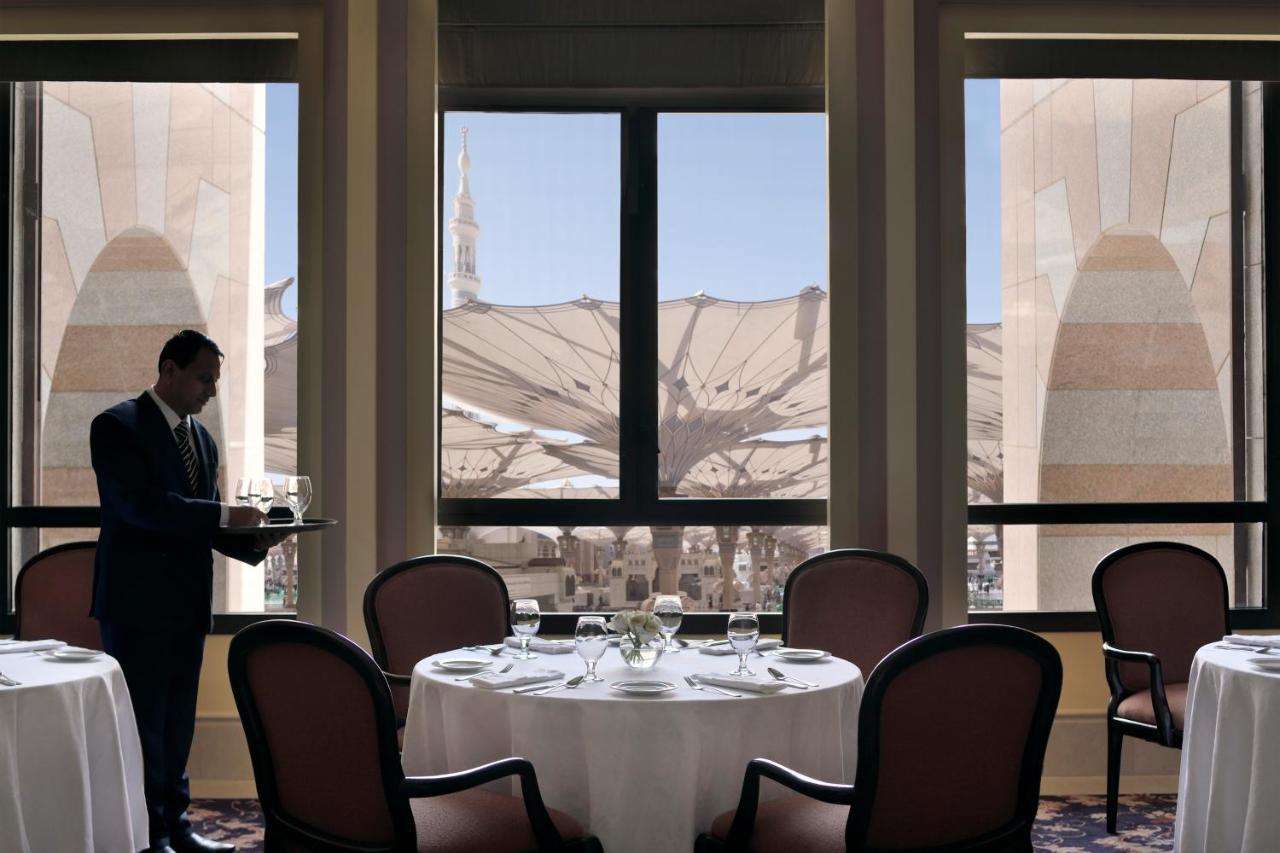 Intercontinental Dar Al Iman Madinah By Ihg Hotel Medina Buitenkant foto The photo shows a dining area with elegant decor. A waiter is standing to the side, holding a tray. The tables are neatly set with white tablecloths and fine dining arrangements. Large windows provide a view of an outdoor area featuring architectural