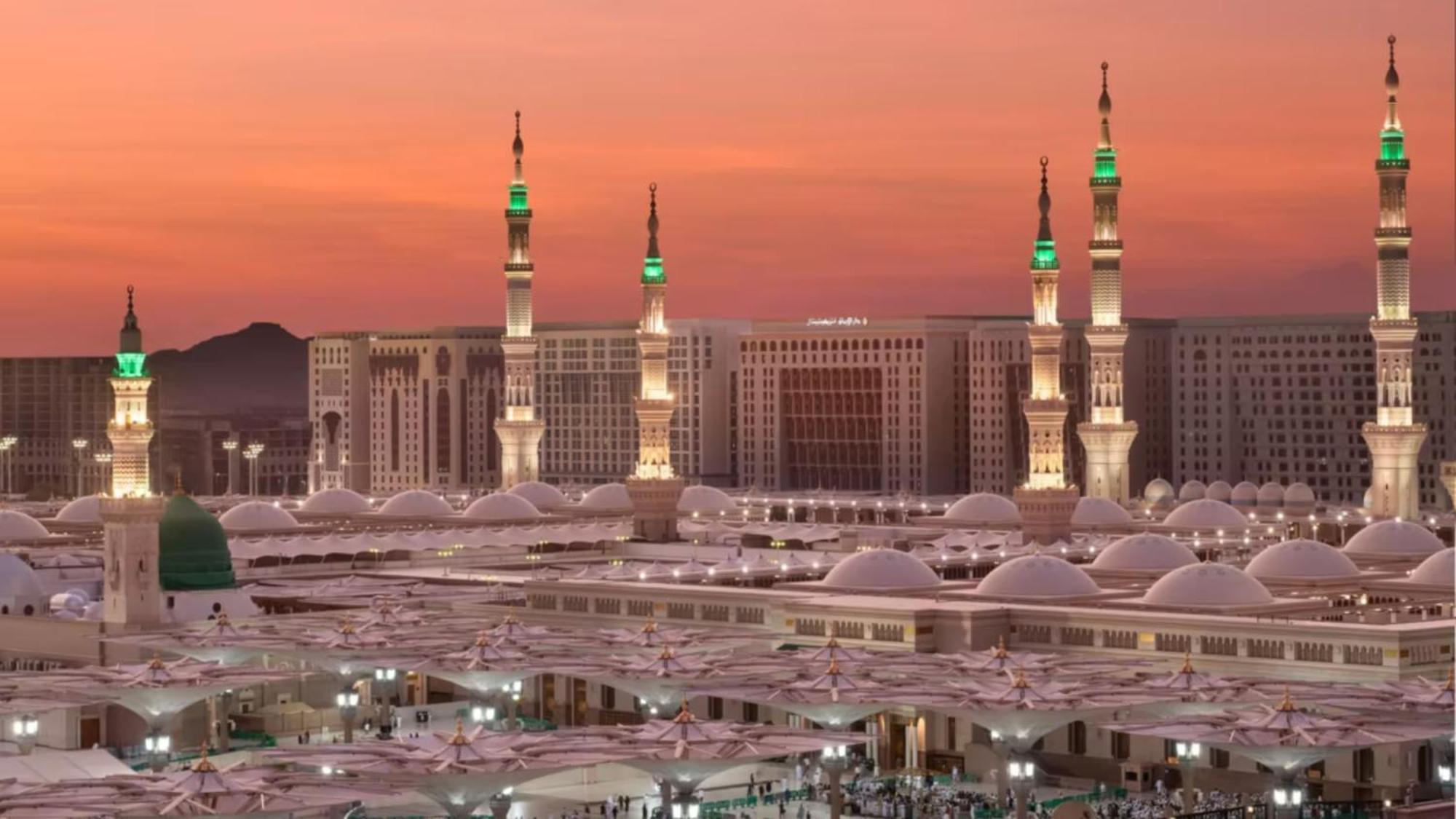 Intercontinental Dar Al Iman Madinah By Ihg Hotel Medina Buitenkant foto The photo depicts a scenic view of a mosque complex, likely during sunset, with several minarets illuminated and showcasing green lights. The architecture features domed roofs, and the setting includes a backdrop of buildings, all bathed in a warm, o