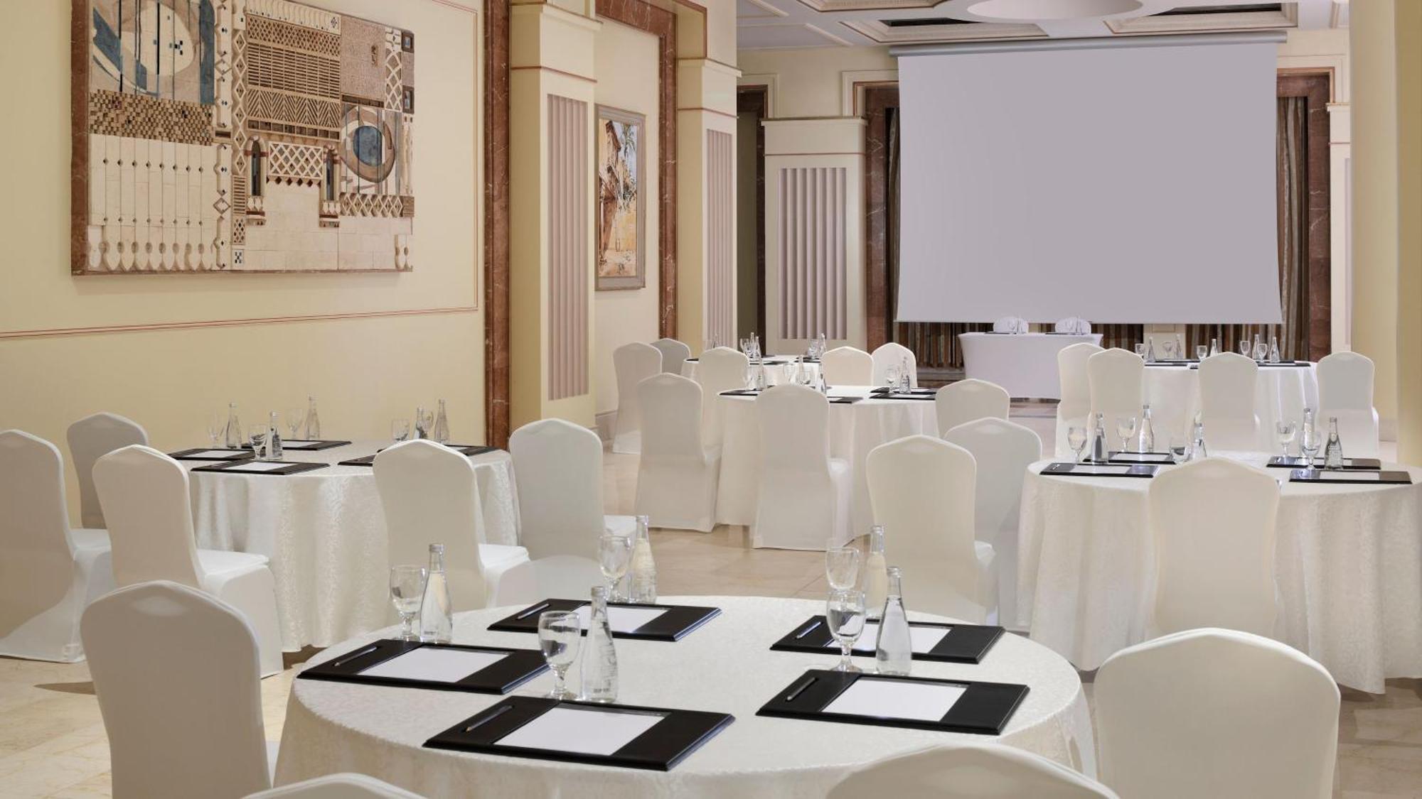 Intercontinental Dar Al Iman Madinah By Ihg Hotel Medina Buitenkant foto The photo depicts an elegantly arranged event space. There are several round tables covered with white tablecloths and surrounded by white chairs. Each table is set with a black folder or menu and a glass of water. The walls are adorned with artworks