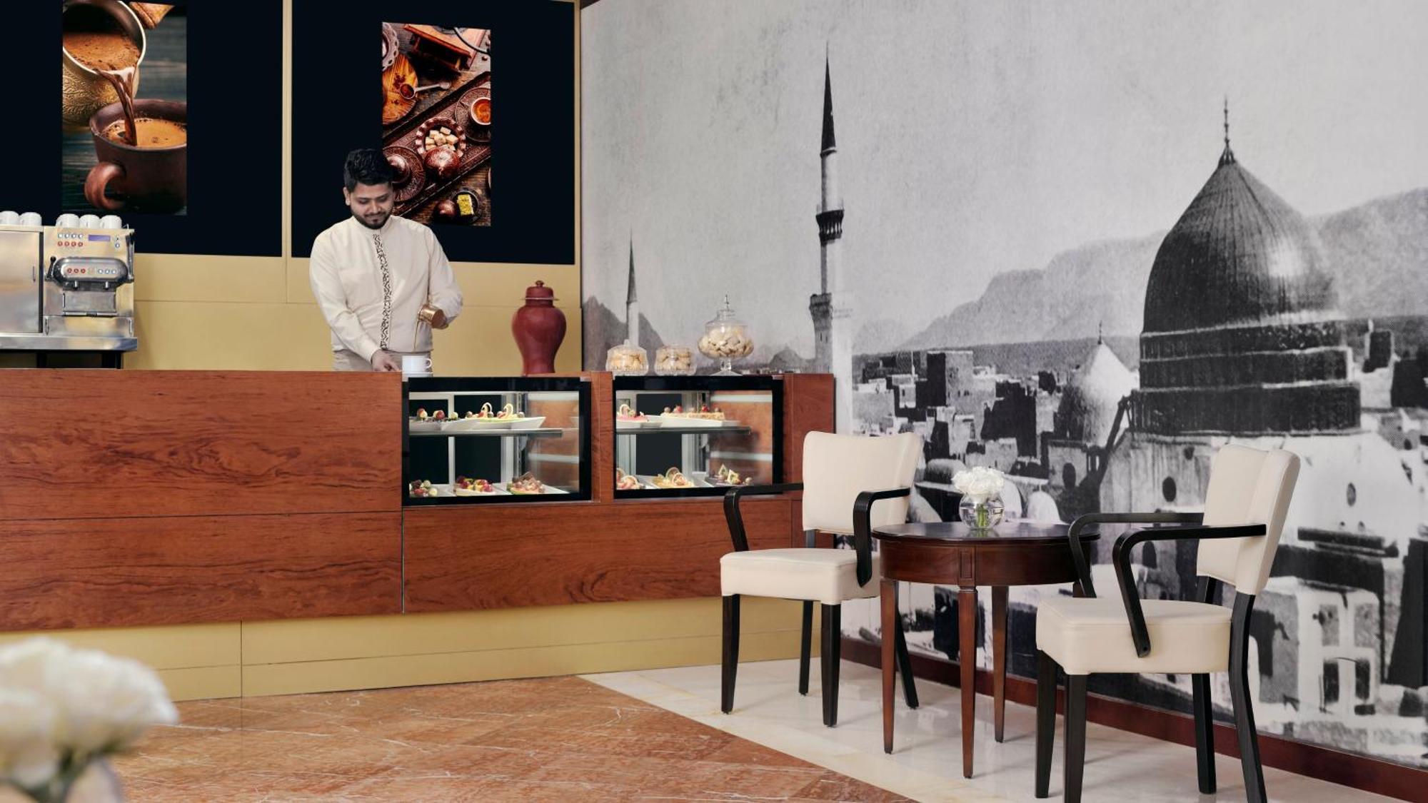 Intercontinental Dar Al Iman Madinah By Ihg Hotel Medina Buitenkant foto The photo shows an elegant cafe or dessert shop interior. A man in traditional attire is standing behind a wooden counter, where various pastries and sweets are displayed. The walls feature a large black-and-white mural of a cityscape, possibly depic
