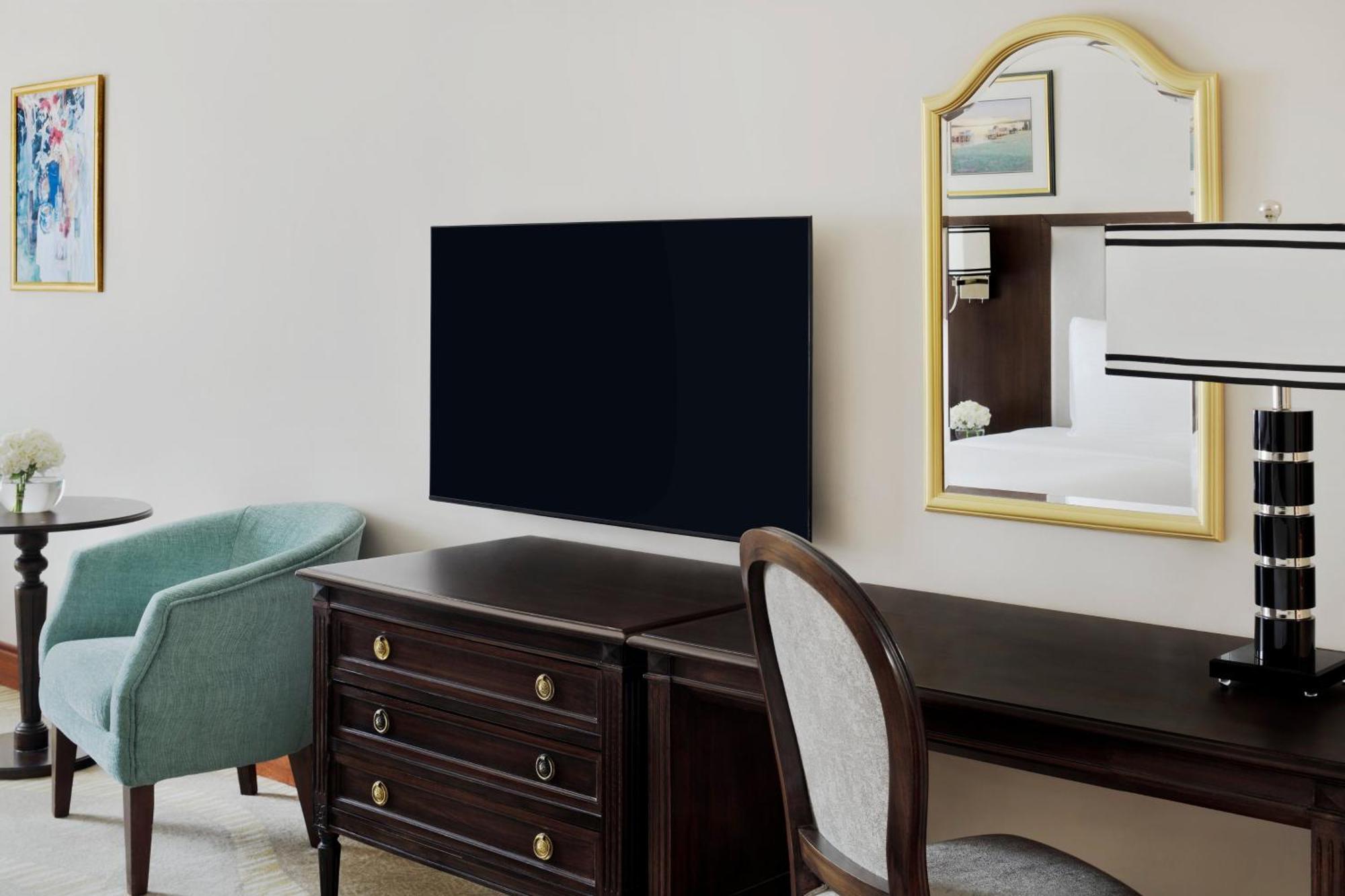 Intercontinental Dar Al Iman Madinah By Ihg Hotel Medina Buitenkant foto The photo shows a hotel room interior featuring a mounted flat-screen television on the wall. Below the TV, there is a dark wooden dresser with drawers. To the right, there's a wooden desk and a chair with a light-colored seat. A small table with a l
