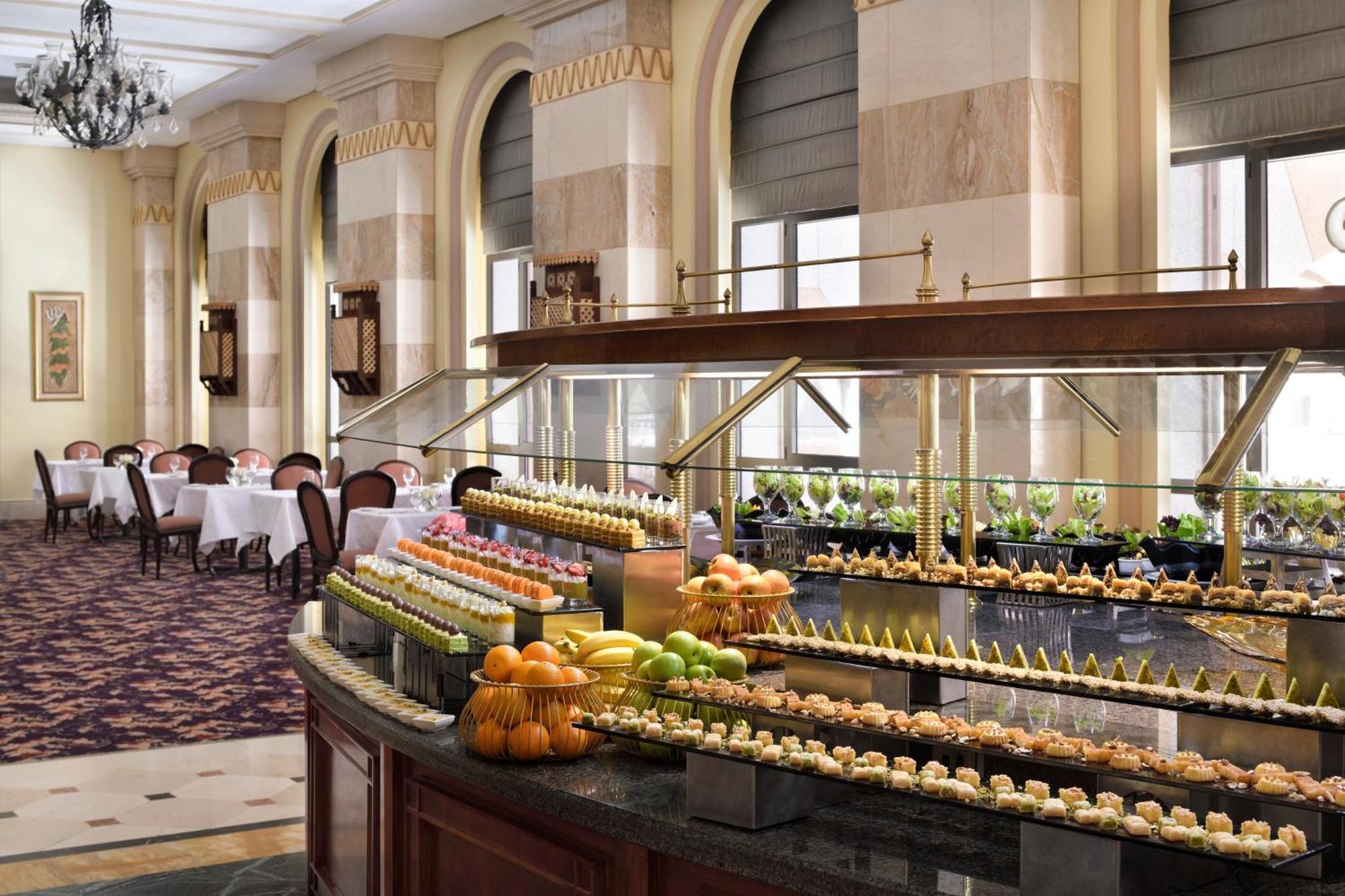 Intercontinental Dar Al Iman Madinah By Ihg Hotel Medina Buitenkant foto The photo depicts an elegant dining area featuring a buffet-style display. In the foreground, a beautifully arranged buffet counter showcases an array of desserts, including miniature pastries and colorful fruit. The setting has a sophisticated ambia