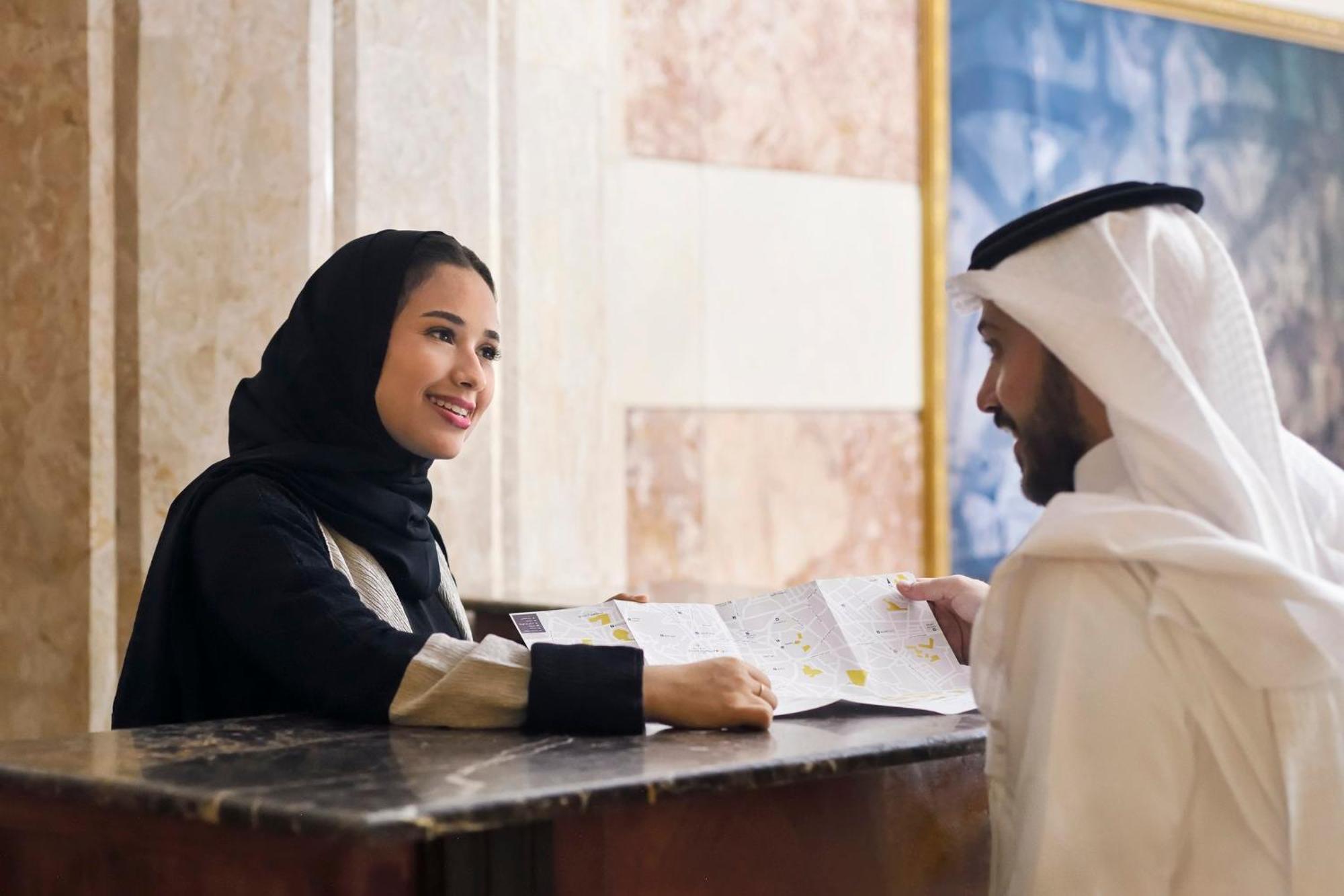 Intercontinental Dar Al Iman Madinah By Ihg Hotel Medina Buitenkant foto The photo depicts a woman in a black hijab and a light-colored outfit smiling as she interacts with a man wearing traditional Emirati attire, including a white kandura and ghutrah (headscarf). They are at a reception desk in what appears to be a hote