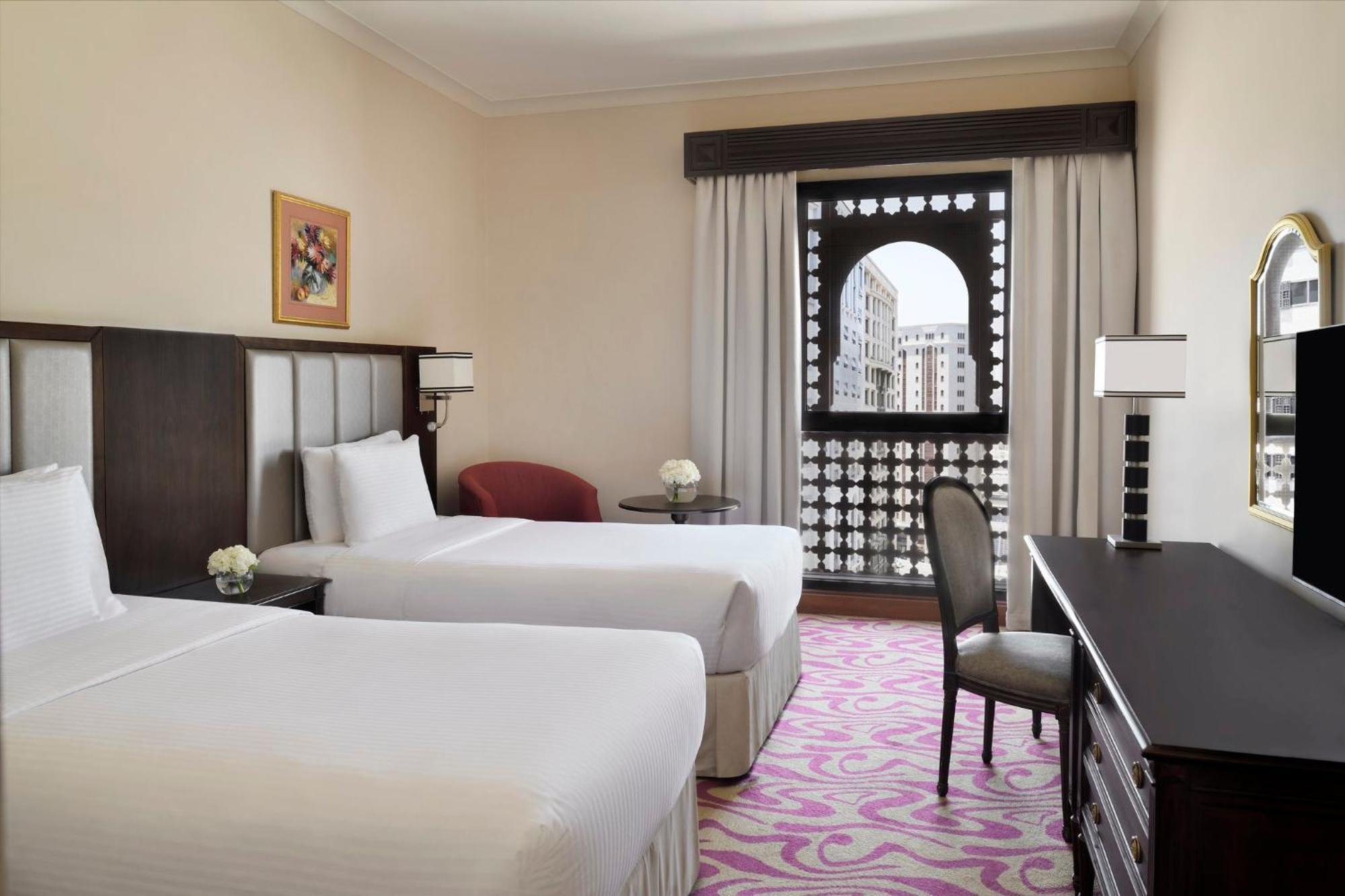Intercontinental Dar Al Iman Madinah By Ihg Hotel Medina Buitenkant foto The photo shows a hotel room featuring two single beds with white bedding. The room has a light, neutral color scheme and is decorated with minimal furnishings. There's a small seating area with a red chair and a round table, as well as a desk with a