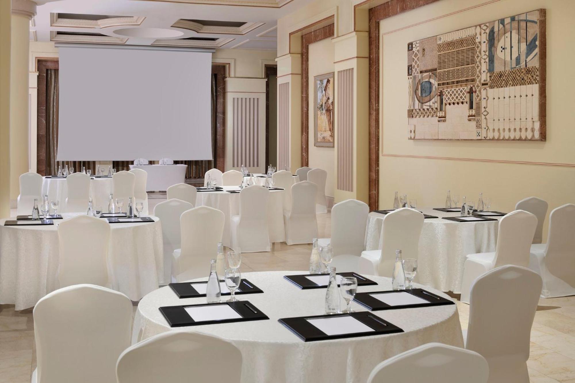 Intercontinental Dar Al Iman Madinah By Ihg Hotel Medina Buitenkant foto The photo shows a well-decorated event space or meeting room. It features several round tables covered with white tablecloths and surrounded by white chairs. Each table has black placemats and glass water bottles, suggesting a formal setup for a gath