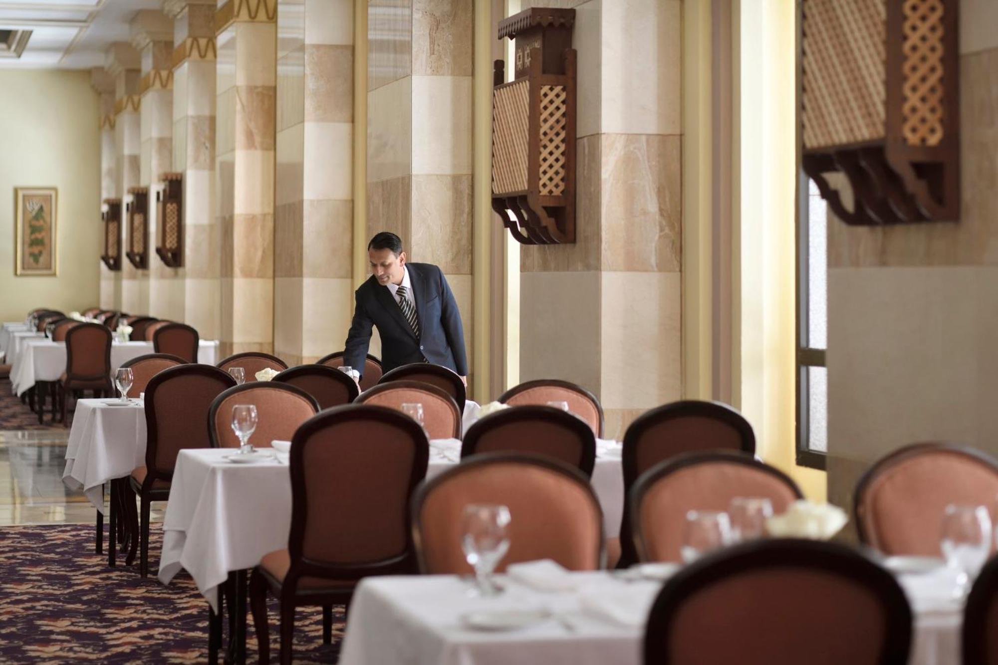 Intercontinental Dar Al Iman Madinah By Ihg Hotel Medina Buitenkant foto The photo depicts a dining area in an elegant restaurant. There are several tables set with white tablecloths, each accompanied by chairs. A waiter, dressed in a formal suit, is seen arranging or tending to one of the tables. The restaurant features 
