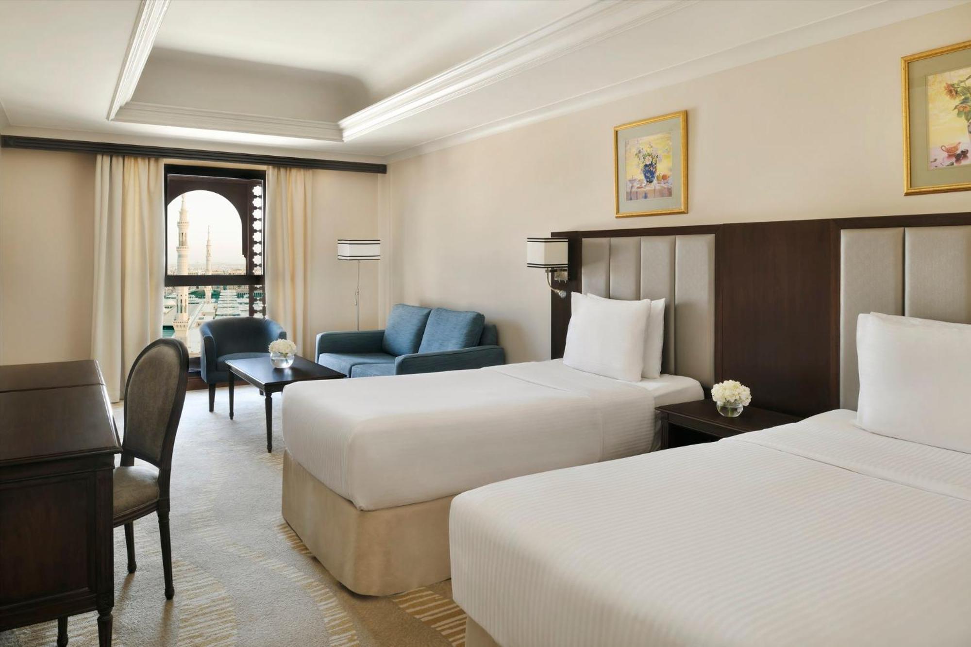 Intercontinental Dar Al Iman Madinah By Ihg Hotel Medina Buitenkant foto The photo shows a well-appointed hotel room featuring two twin beds with white bedding. There is a small desk with a chair, a comfortable blue sofa, and a coffee table in the sitting area. The room has a warm, neutral color scheme, accented by two pa