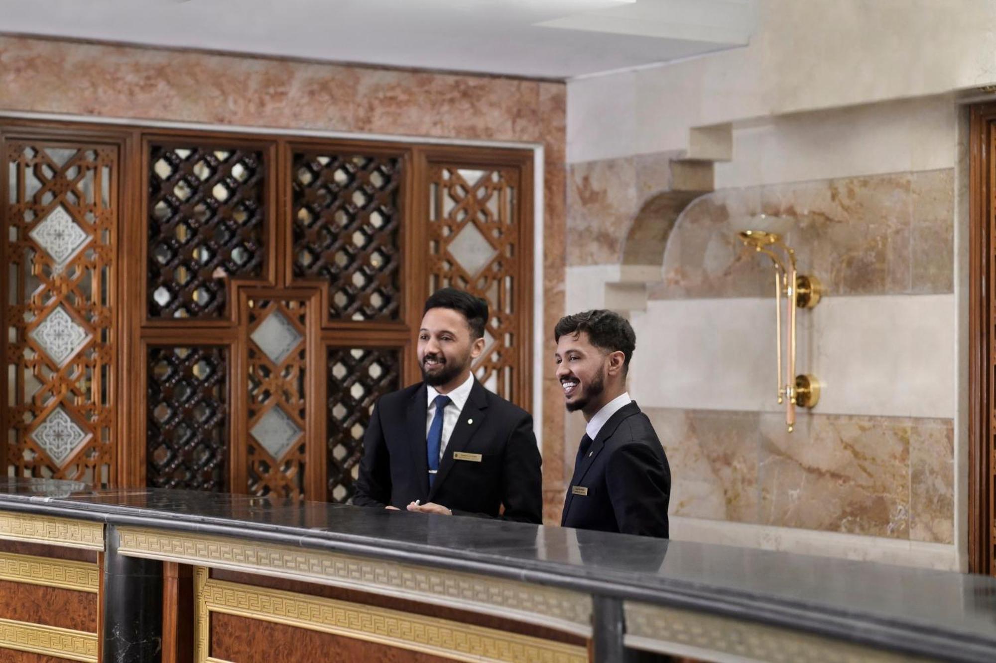 Intercontinental Dar Al Iman Madinah By Ihg Hotel Medina Buitenkant foto The photo shows two men standing behind a reception desk in an elegant setting, likely a hotel or similar establishment. They are both dressed in formal attire, wearing suits and ties, and appear to be smiling or engaging in conversation. The backgro