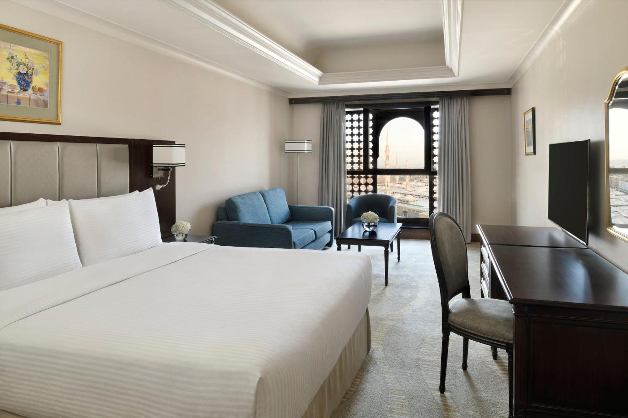 Intercontinental Dar Al Iman Madinah By Ihg Hotel Medina Buitenkant foto The photo shows a hotel room designed with a modern and comfortable aesthetic. It features a large bed with white linens, a seating area with a blue sofa, and a small table. There is a wooden desk with a chair and a television opposite the bed. Natur
