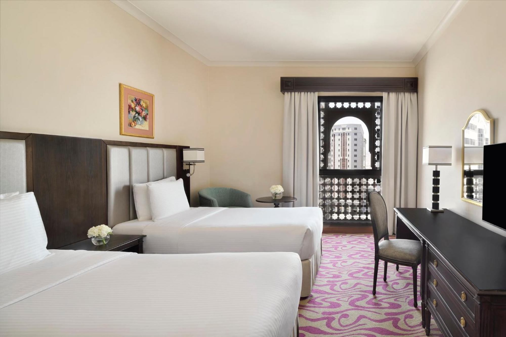 Intercontinental Dar Al Iman Madinah By Ihg Hotel Medina Buitenkant foto The photo shows a hotel room featuring two twin beds with white bedding. There is a decorative piece of art on the wall above one of the beds. A small round table with a flower arrangement is placed next to the beds. A desk and chair are situated aga