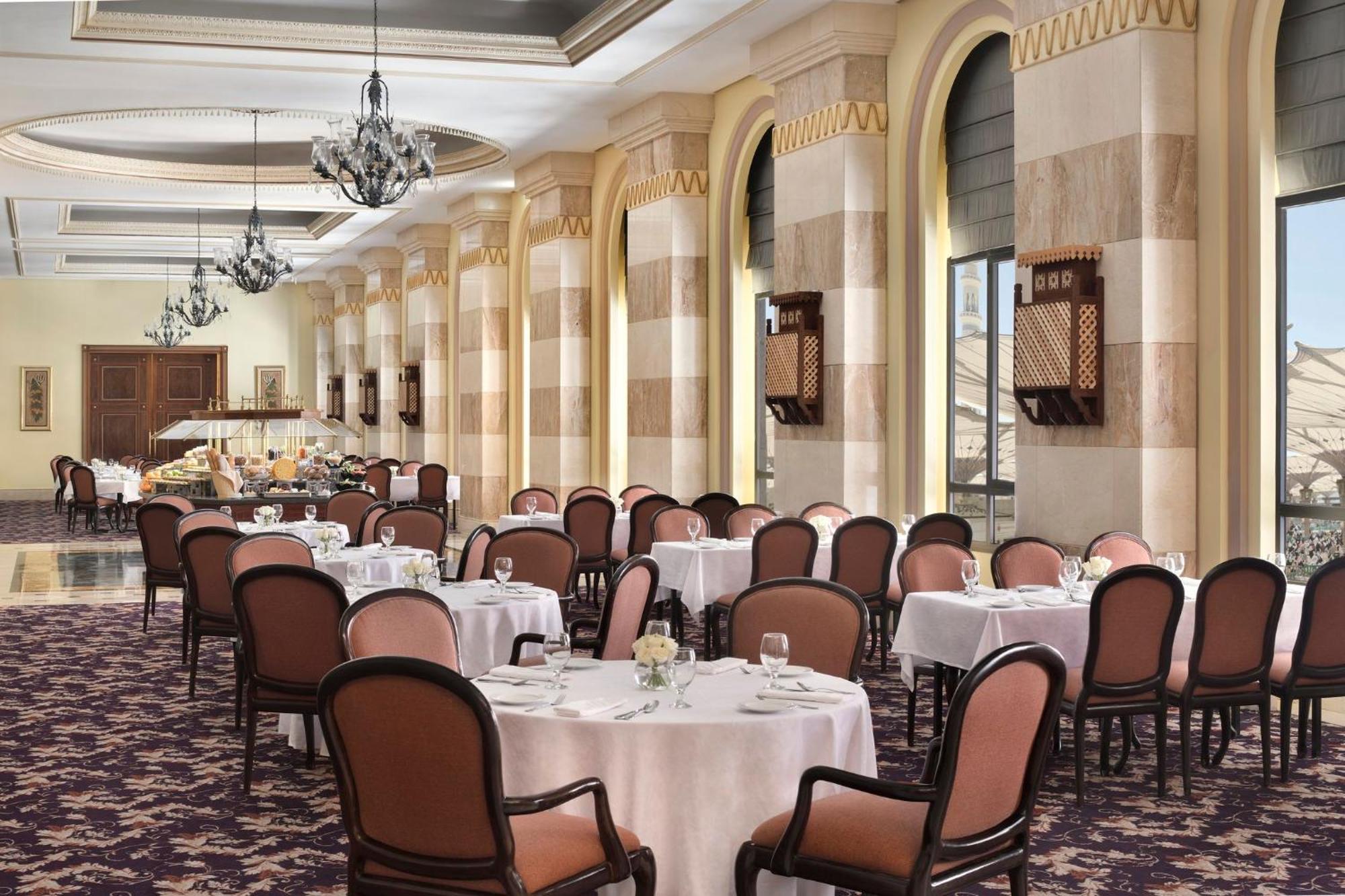 Intercontinental Dar Al Iman Madinah By Ihg Hotel Medina Buitenkant foto The photo depicts an elegant dining room setting. It features several round tables covered with white tablecloths, each set with dishes and glassware. The room has a sophisticated ambiance, characterized by high ceilings adorned with chandeliers and 