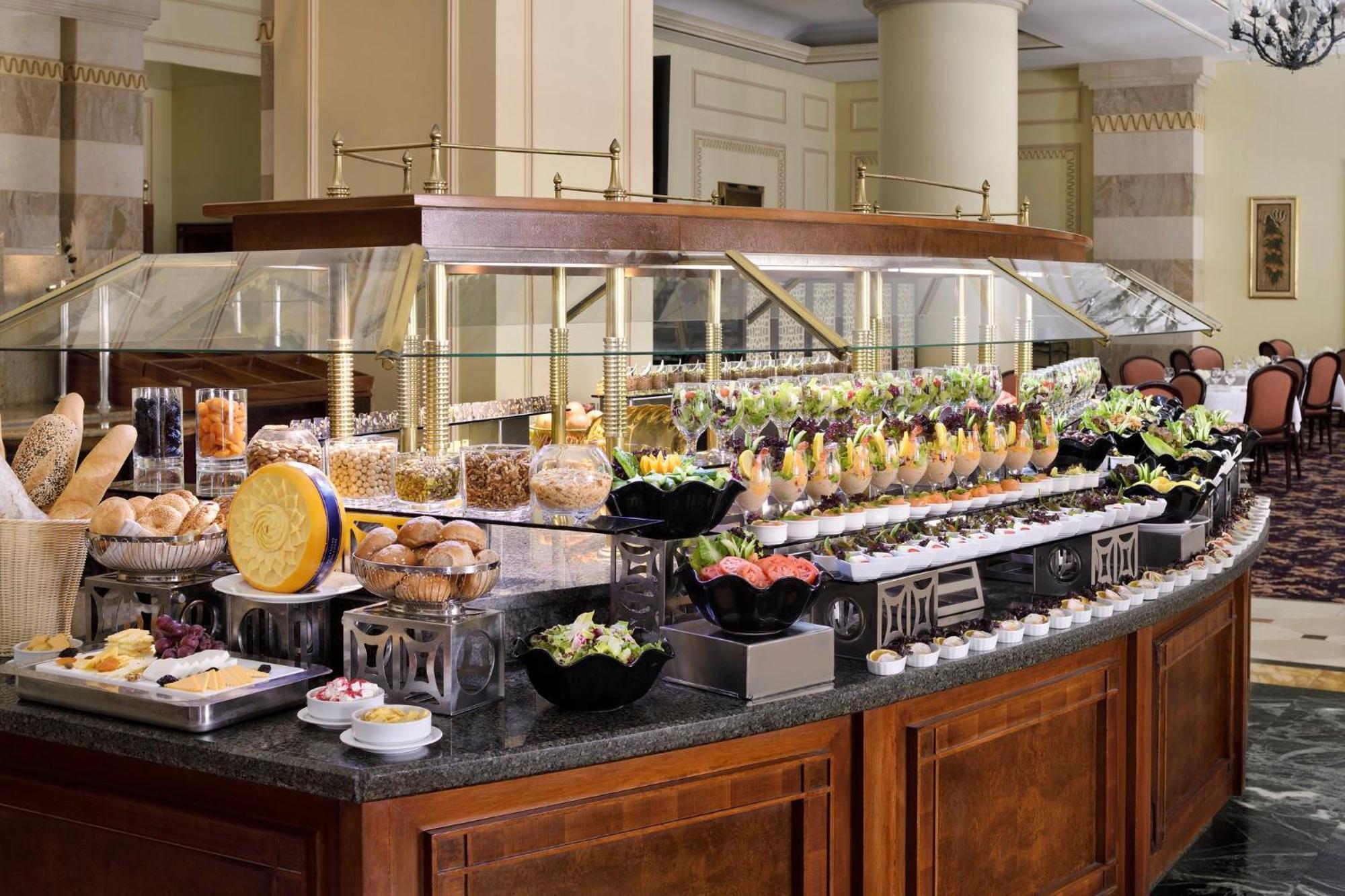 Intercontinental Dar Al Iman Madinah By Ihg Hotel Medina Buitenkant foto The photo shows an elegantly arranged buffet with a variety of food items. There is a large, curved wooden display with glass covers, showcasing an assortment of dishes. These include different types of breads and rolls, salads, garnishes, and desser