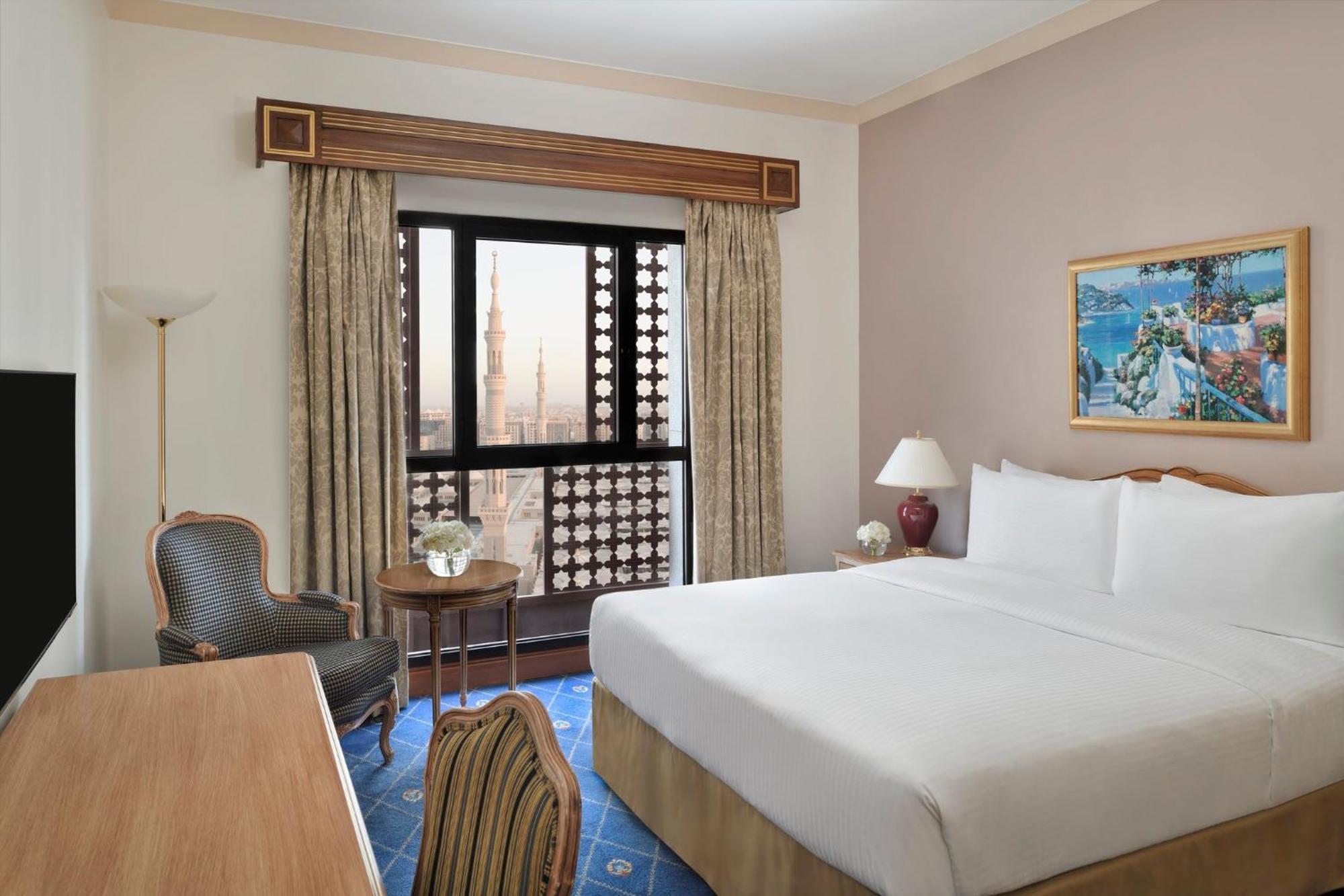 Intercontinental Dar Al Iman Madinah By Ihg Hotel Medina Buitenkant foto The photo shows a hotel room with a modern design. There is a large bed with white linens placed against the wall. Next to the bed, there is a small bedside table with a lamp. A comfortable armchair is situated nearby, accompanied by a wooden desk. T