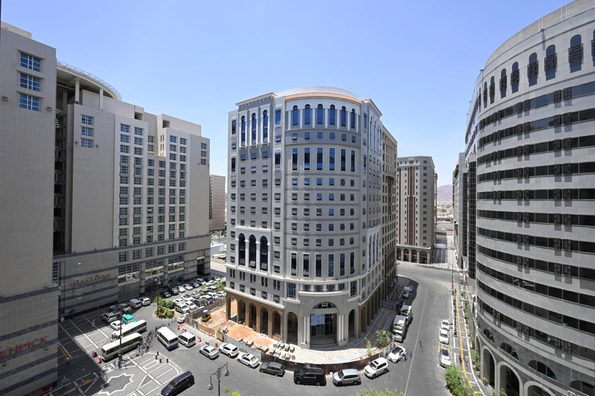 Intercontinental Dar Al Iman Madinah By Ihg Hotel Medina Buitenkant foto The photo shows a modern urban scene featuring several buildings, including a prominent rounded building with a glass façade. This building appears to be situated at an intersection, surrounded by other high-rise structures that have distinct archite