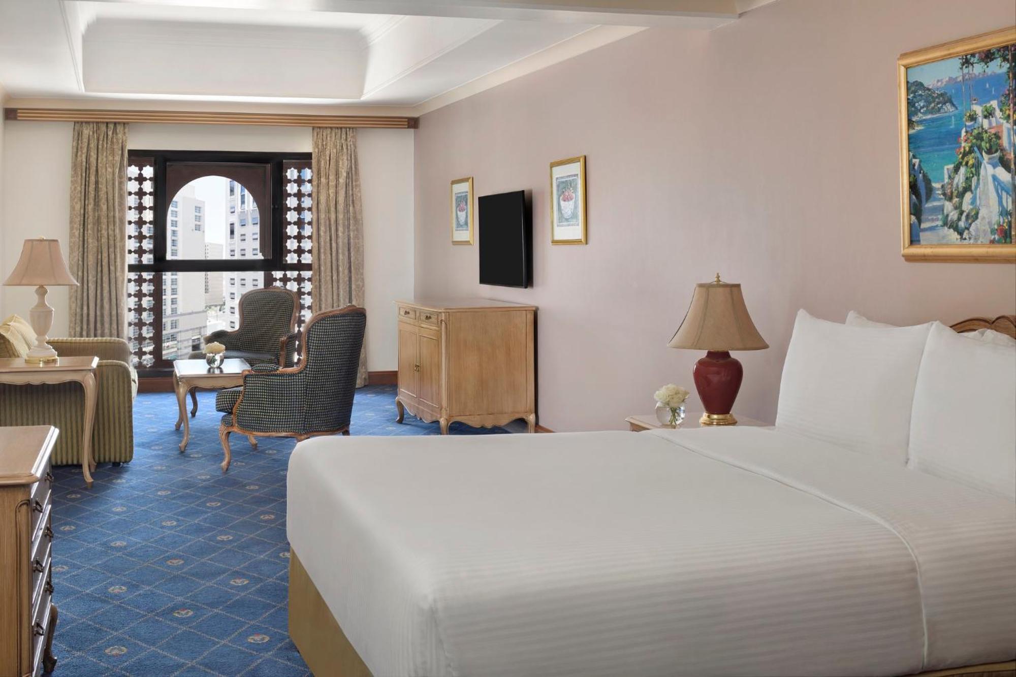 Intercontinental Dar Al Iman Madinah By Ihg Hotel Medina Buitenkant foto The photo shows a well-furnished hotel room. It features a large bed with a white duvet, two bedside lamps, and decorative paintings on the walls. There is a comfortable seating area with two chairs and a small table. A wooden dresser is positioned a