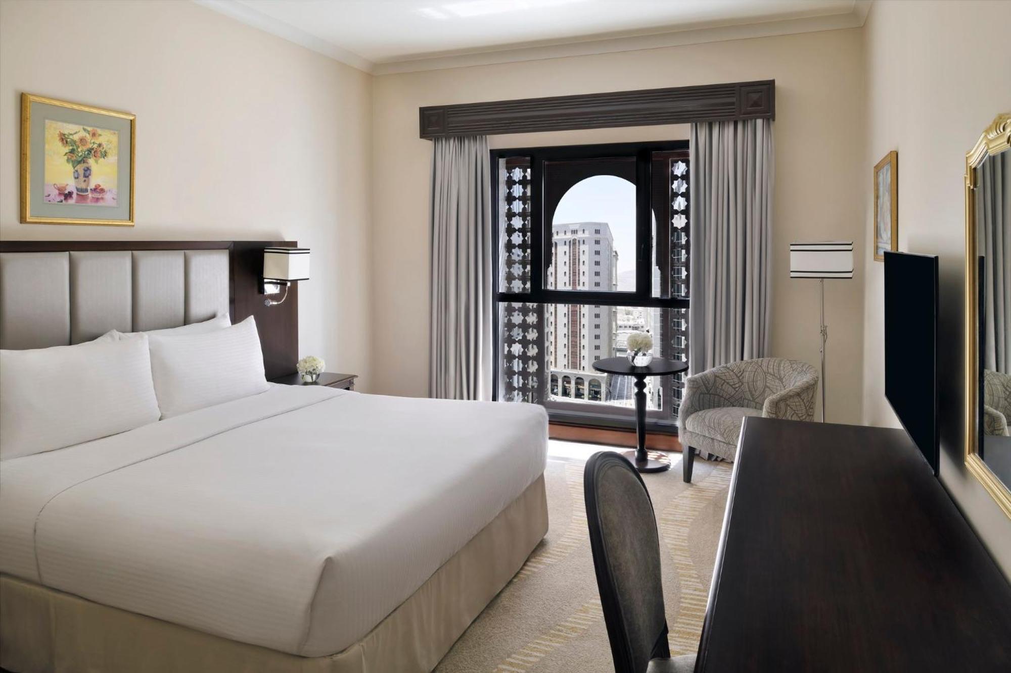 Intercontinental Dar Al Iman Madinah By Ihg Hotel Medina Buitenkant foto The photo shows a hotel room with a modern and minimalistic design. It features a large bed with white linens, a side table, and a comfortable chair near a window. The window has a view of a cityscape, and elegant curtains frame the window. There is 