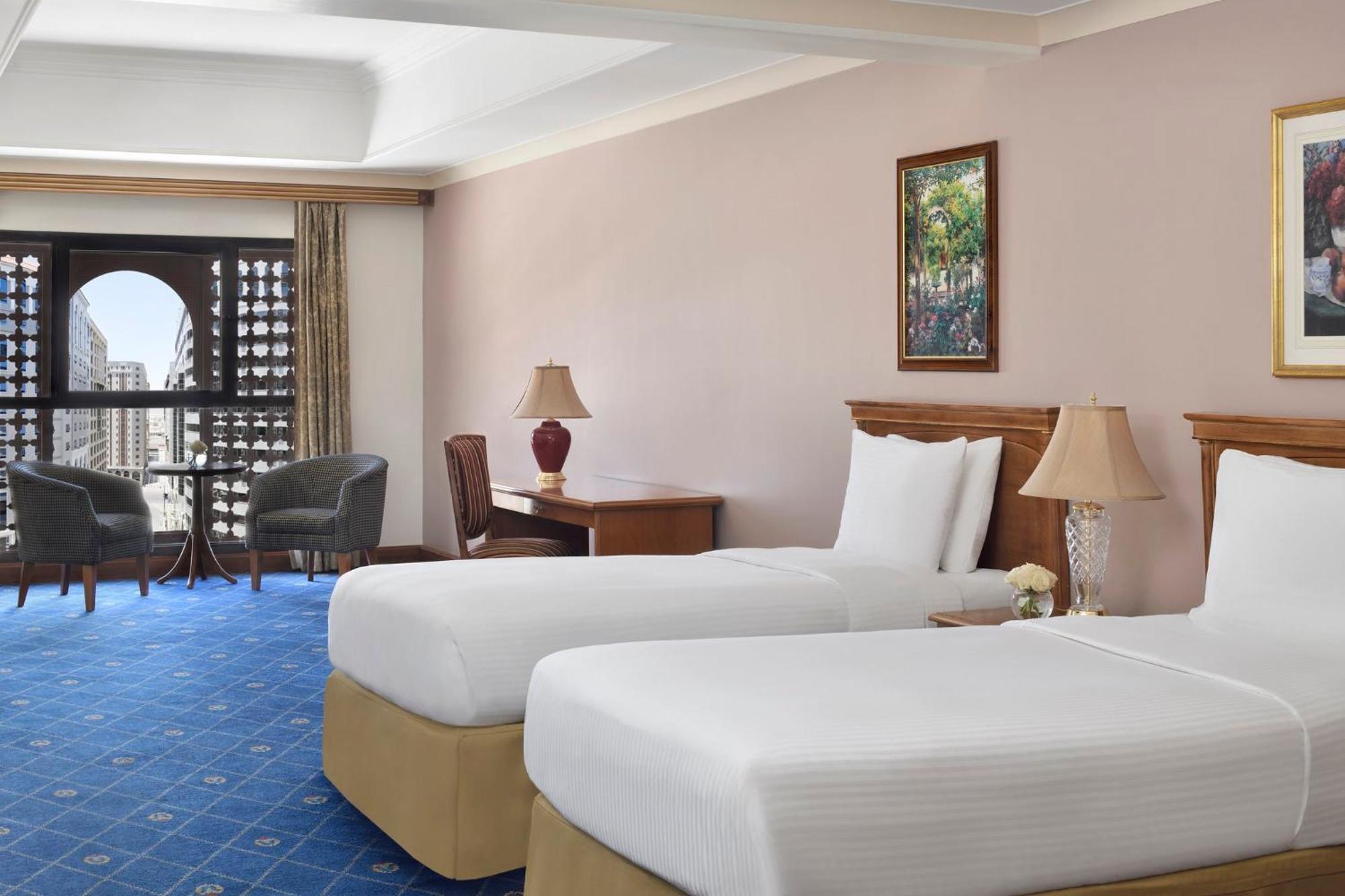 Intercontinental Dar Al Iman Madinah By Ihg Hotel Medina Buitenkant foto The photo shows a nicely designed hotel room featuring two twin beds with white linens and beige bases. In the background, there's a wooden desk and a lamp, along with a pair of comfortable armchairs positioned near a large window. The window showcas