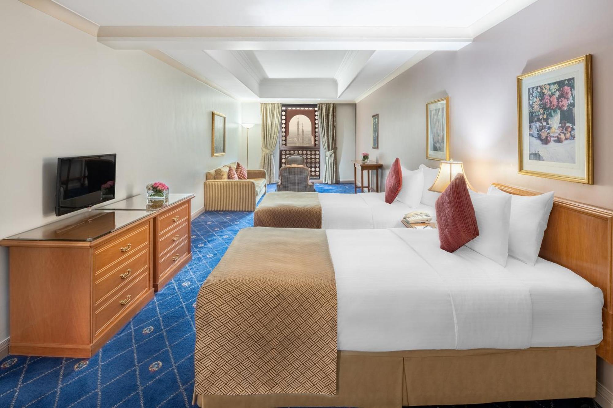 Intercontinental Dar Al Iman Madinah By Ihg Hotel Medina Buitenkant foto The photo shows a hotel room featuring two beds with white linens and decorative pillows. There is a wooden dresser with a television on it. The flooring has a blue carpet with a pattern, and the walls are painted in a light color. The room is well-l