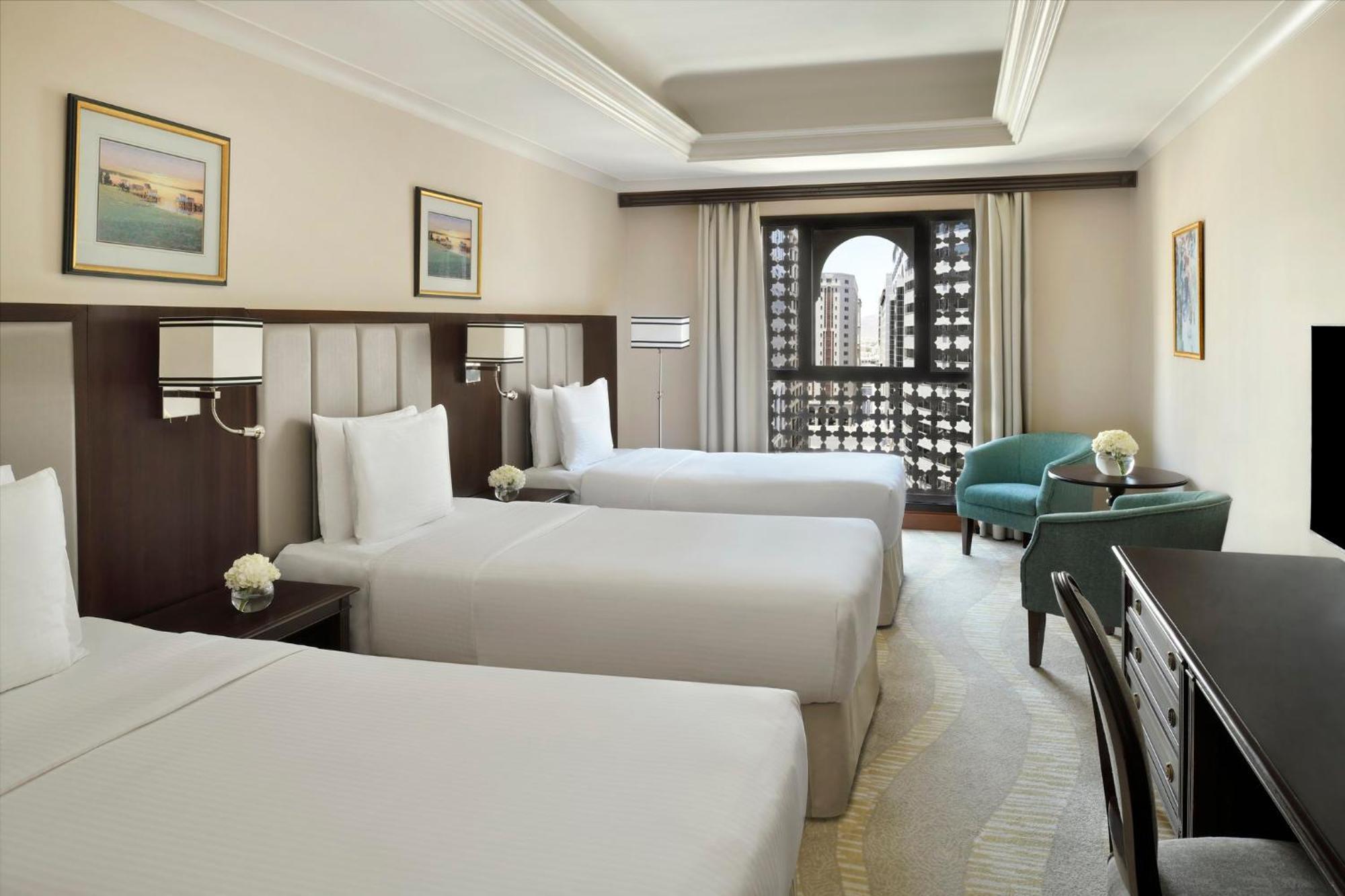 Intercontinental Dar Al Iman Madinah By Ihg Hotel Medina Buitenkant foto The photo shows a hotel room with three single beds, dressed in white linens. There are two paintings on the walls, depicting serene landscapes. A large window allows natural light to enter, and it is adorned with elegant curtains. A smaller seating 