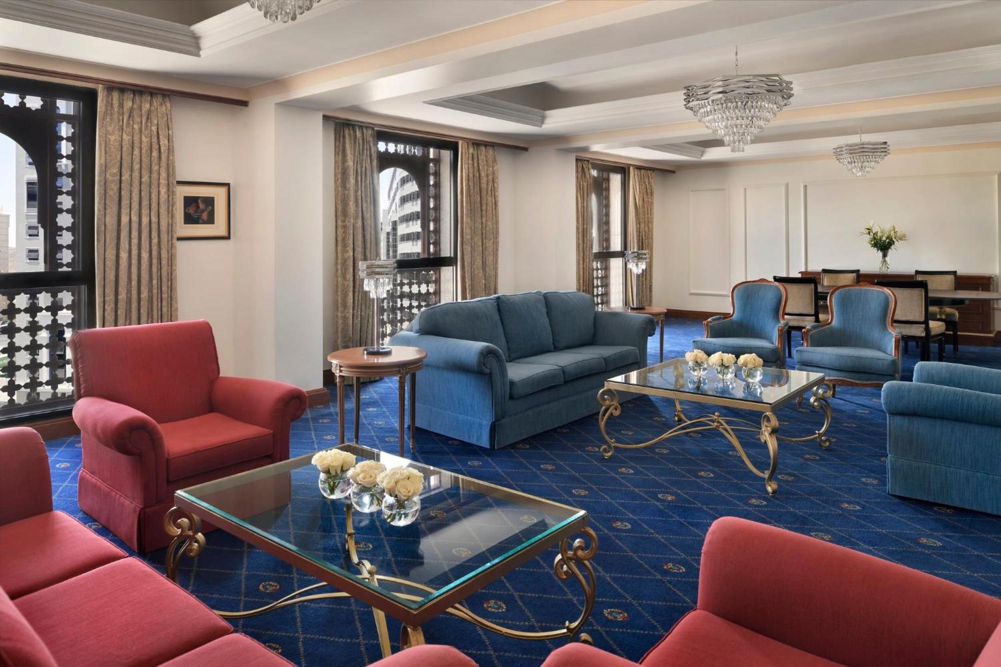 Intercontinental Dar Al Iman Madinah By Ihg Hotel Medina Buitenkant foto The photo shows a luxurious living space or lounge area. It features a combination of furniture, including a blue sofa, several red armchairs, and glass-top coffee tables with gold accents. The flooring is carpeted in a deep blue, and the walls are a