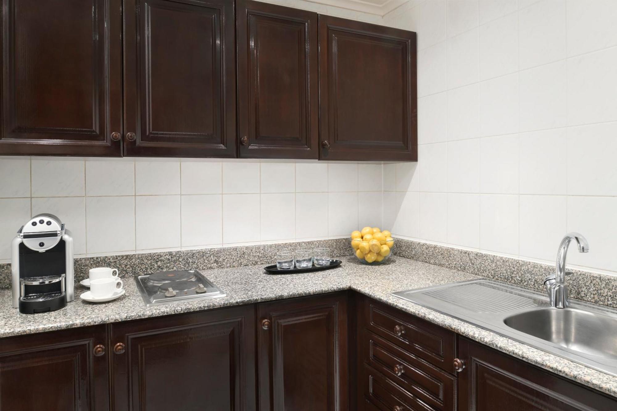 Intercontinental Dar Al Iman Madinah By Ihg Hotel Medina Buitenkant foto The photo shows a kitchen with dark wooden cabinetry and a light-colored countertop. There are a few appliances visible, including a coffee machine and a stovetop with burners. To the right, there is a sink, and a bowl of yellow lemons is placed on t