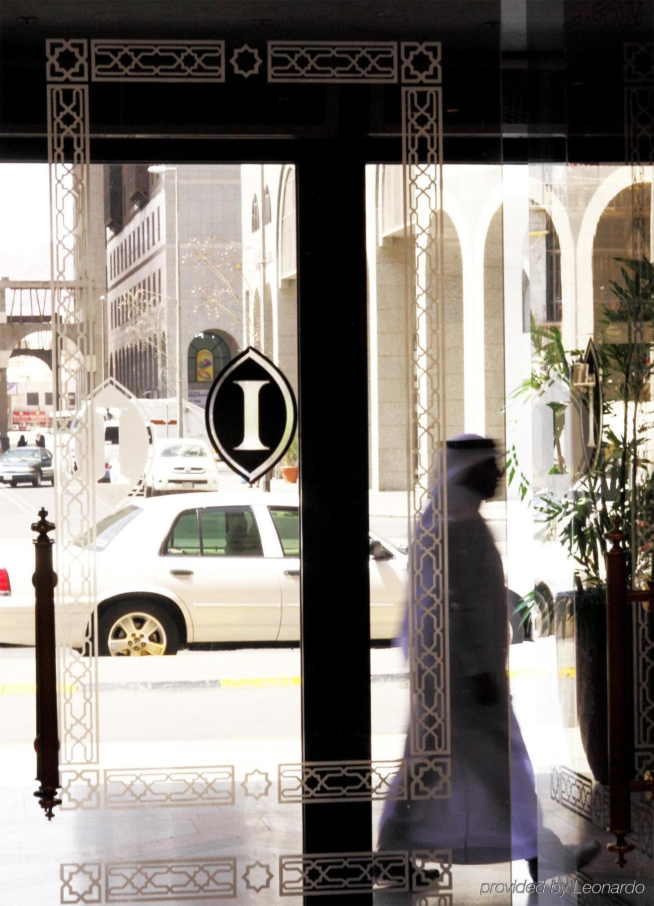 Intercontinental Dar Al Iman Madinah By Ihg Hotel Medina Interieur foto The photo shows a view through a glass door or window, featuring an elegant design with a prominent "I" symbol displayed. Through the glass, there are some parked vehicles visible, including a white car. In the foreground, a shadowy figure of a perso