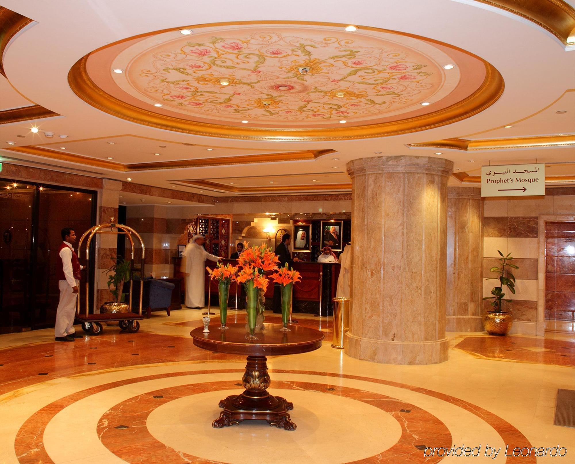 Intercontinental Dar Al Iman Madinah By Ihg Hotel Medina Interieur foto The photo depicts a luxurious hotel lobby. The ceiling features an ornate design with floral patterns, adding elegance to the space. In the center of the lobby, there is a decorative table with a bowl or vase of bright orange flowers. The flooring is