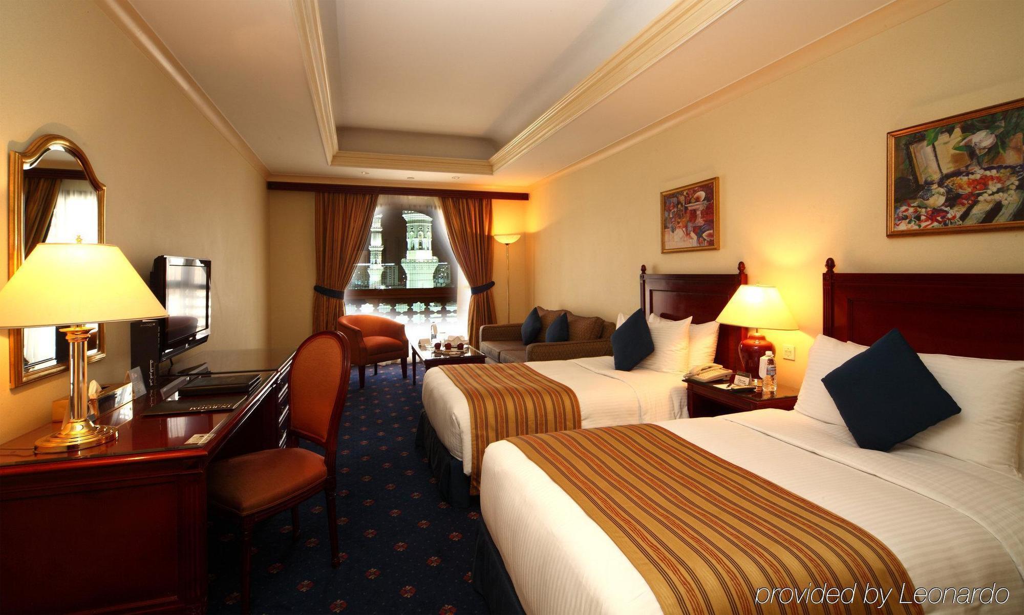 Intercontinental Dar Al Iman Madinah By Ihg Hotel Medina Kamer foto The photo shows a hotel room featuring two beds with white linens and brown striped throws. There is a wooden desk and chair, alongside a television. The walls are painted in a warm tone, and there are decorative paintings. Large windows with curtain