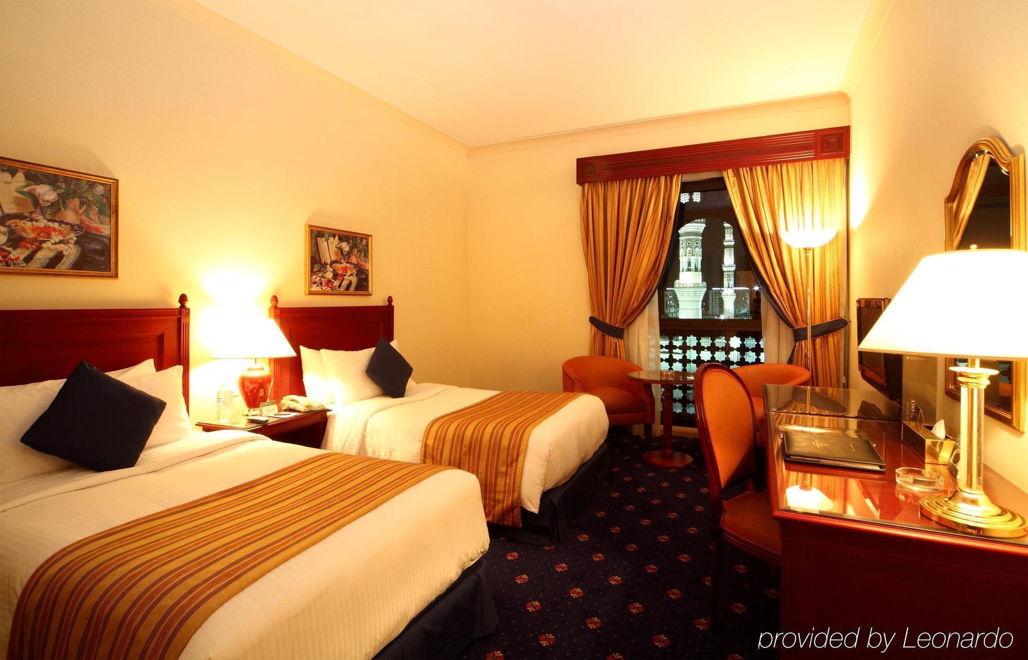 Intercontinental Dar Al Iman Madinah By Ihg Hotel Medina Kamer foto The photo shows a hotel room featuring two twin beds with white linens and brown striped bedspreads. There are decorative pillows on the beds, and the room has warm-colored walls. To the side, there's a small sitting area with a round table and a cha