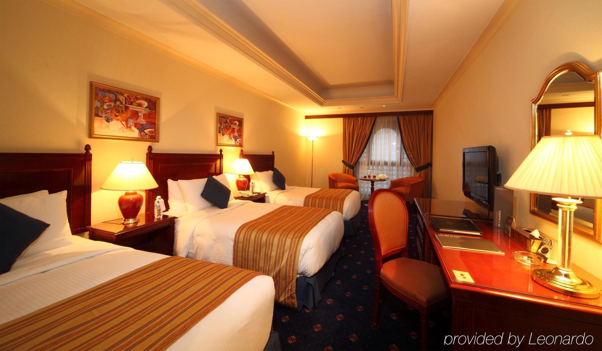 Intercontinental Dar Al Iman Madinah By Ihg Hotel Medina Kamer foto The photo shows a hotel room with a warm and inviting atmosphere. There are two beds with white and gold bedding, complemented by decorative pillows. Each bed has a bedside lamp. In the background, there's a small seating area with a couple of chairs