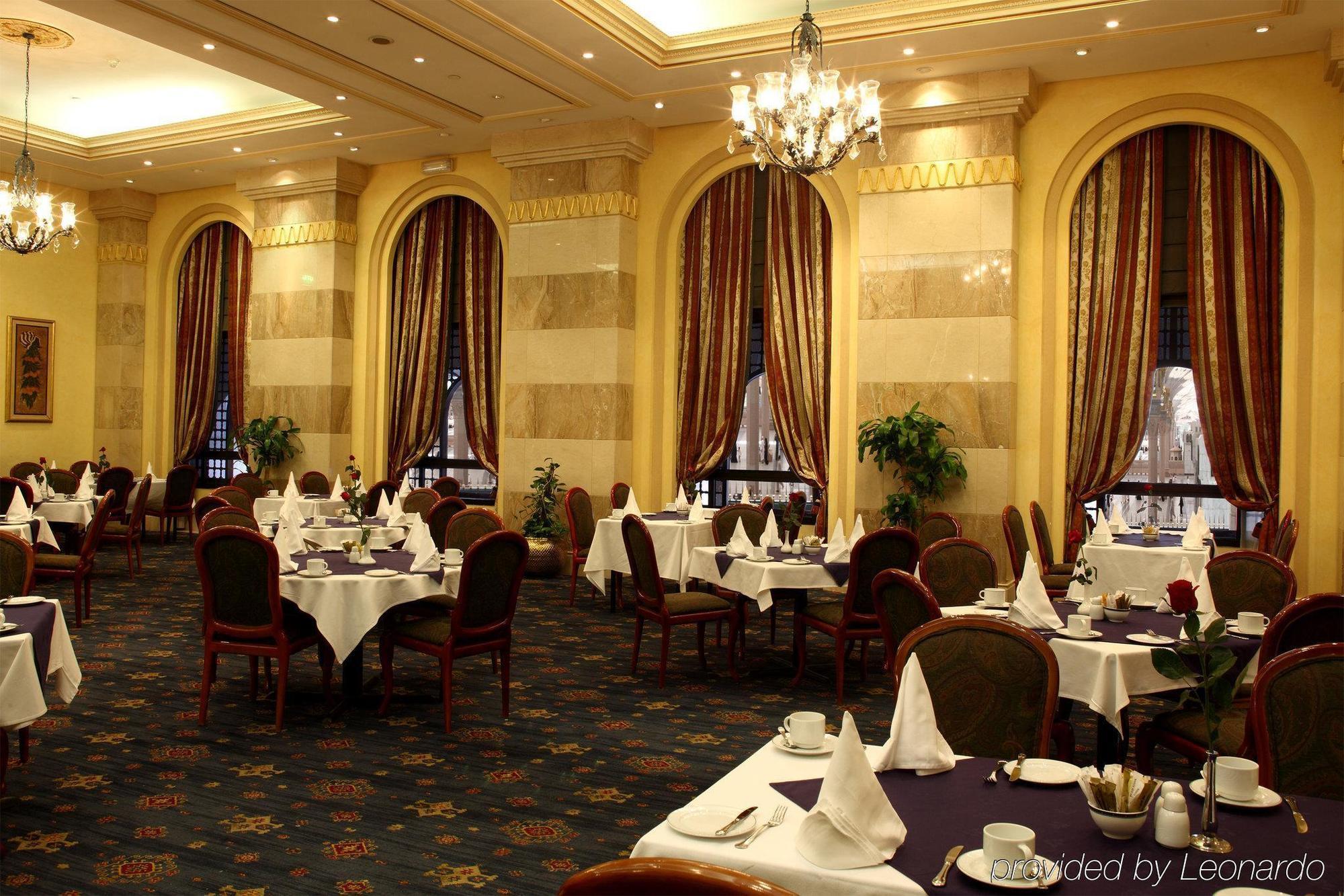 Intercontinental Dar Al Iman Madinah By Ihg Hotel Medina Restaurant foto The photo depicts an elegant restaurant interior. The space features ornate decor with tall windows draped in rich, patterned curtains. Tables are neatly set with white tablecloths and napkins, and there are plush chairs arranged around them. The flo