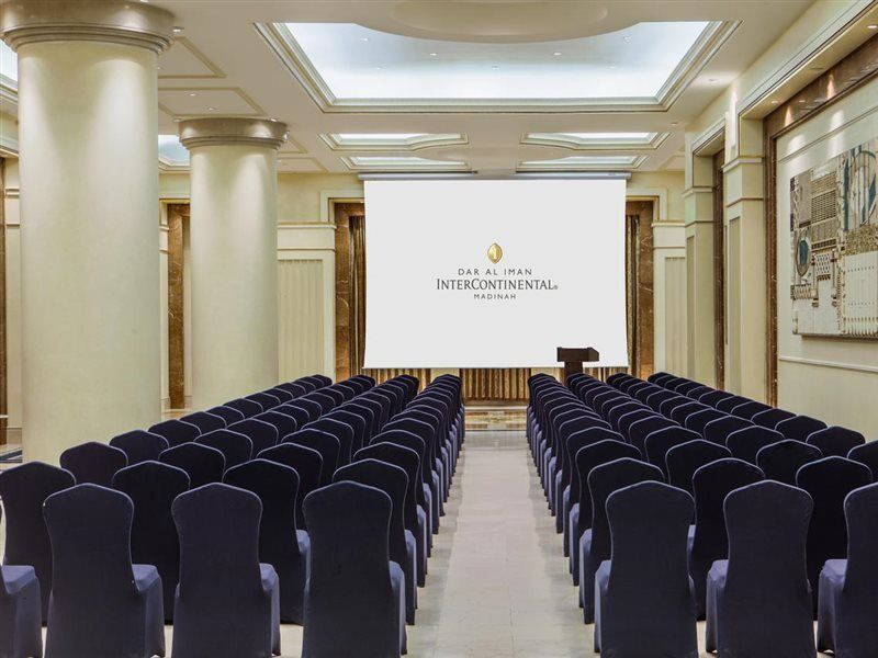 Intercontinental Dar Al Iman Madinah By Ihg Hotel Medina Buitenkant foto The photo shows a conference or meeting room set up. There are rows of dark blue chairs arranged facing a large screen at the front of the room. The screen displays the logo of the Intercontinental hotel, indicating that this space is likely intended