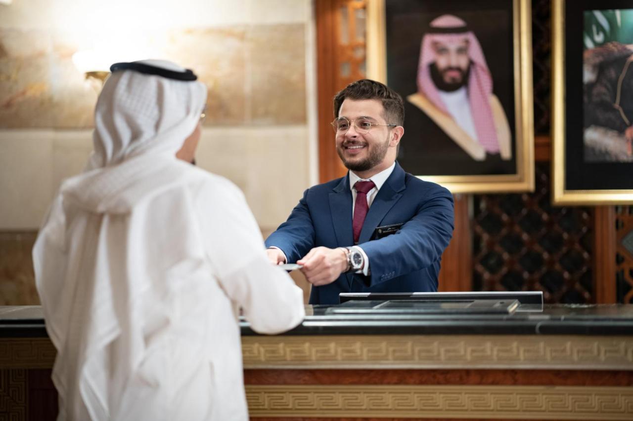 Intercontinental Dar Al Iman Madinah By Ihg Hotel Medina Buitenkant foto The photo shows a hotel reception area. A male receptionist, dressed in a navy suit with a tie, is smiling and handing an item to a guest who is wearing traditional white attire, possibly a kandura or thobe. The background features elegantly decorate