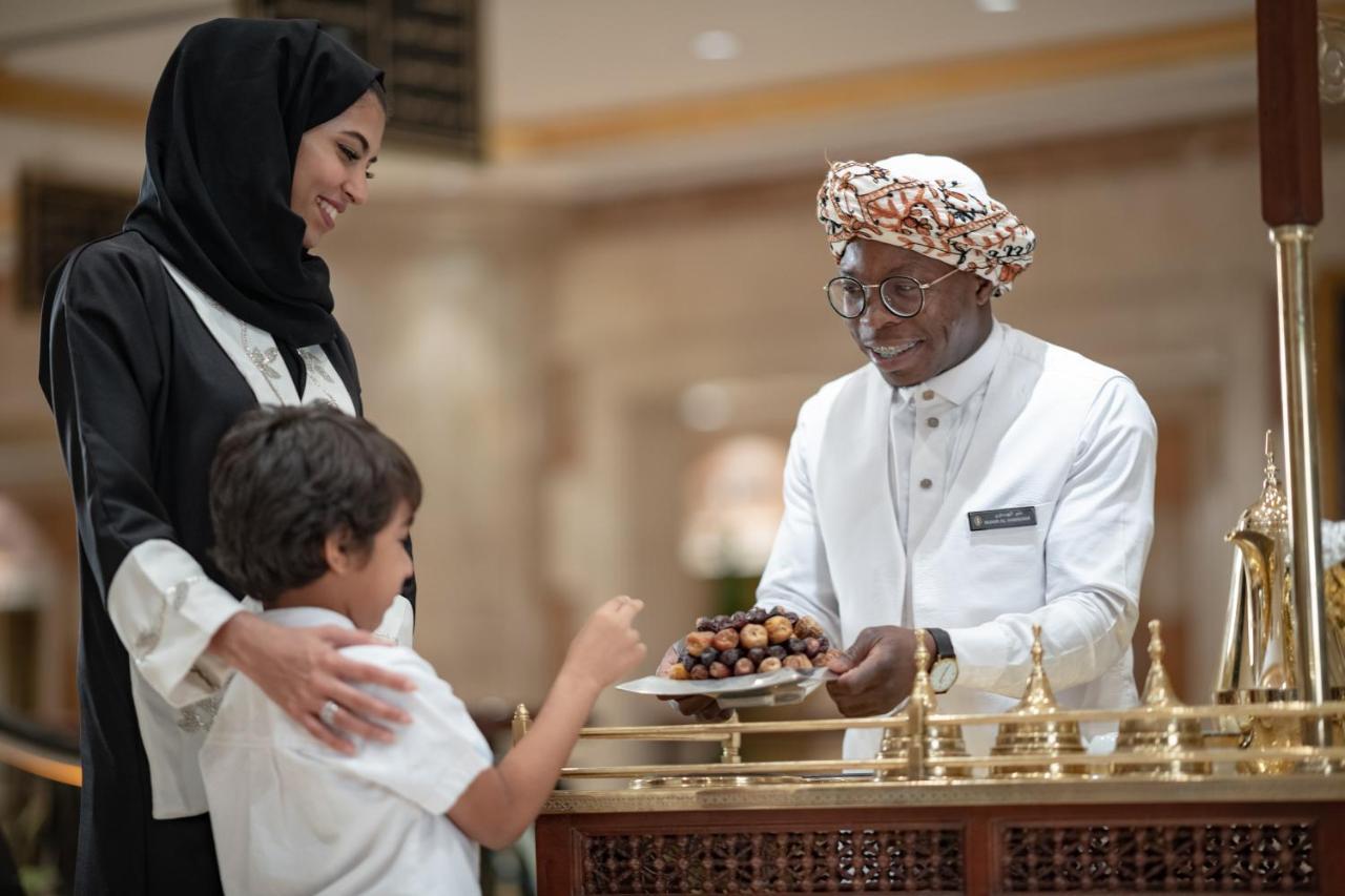 Intercontinental Dar Al Iman Madinah By Ihg Hotel Medina Buitenkant foto The photo depicts a warm interaction between three people. A woman, dressed in a traditional black attire and a headscarf, stands beside a young boy who is reaching out towards a plate of dates held by a smiling man in a white traditional outfit with